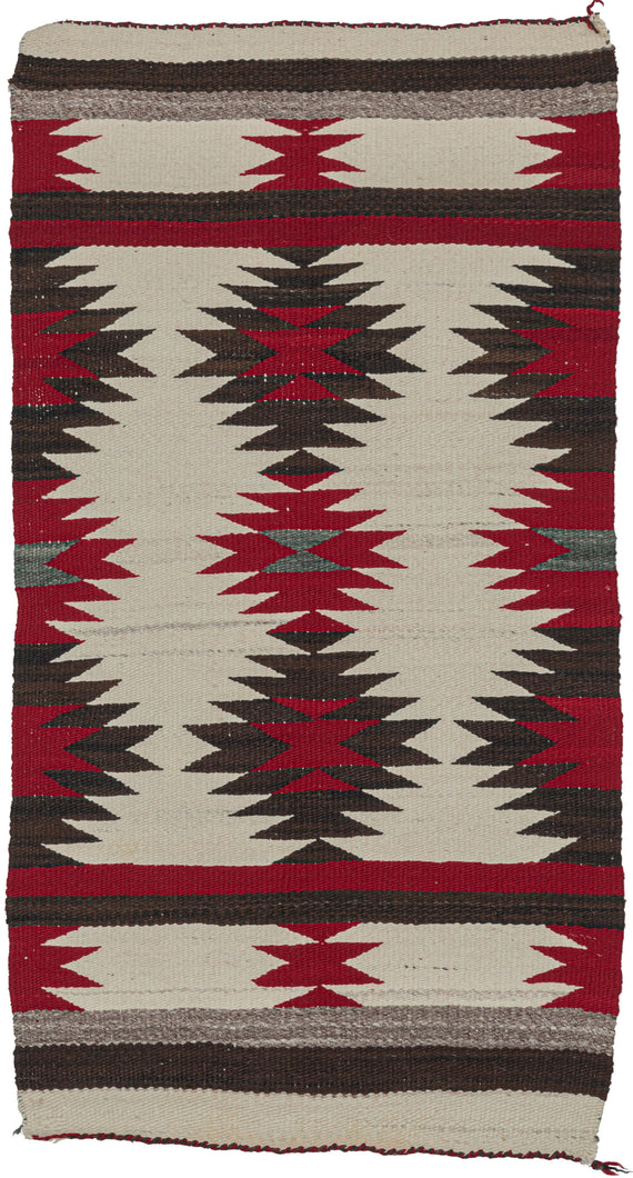 This Navajo rug was handwoven in the southwest United States in the middle of the 20th century.  It features a repeating sunburst pattern in brown, red, and gray on an ivory ground. The pattern features two horizontal bands of solid red and brown in the interior and is finished with solid horizontal bands of gray, brown, and ivory on both ends. 