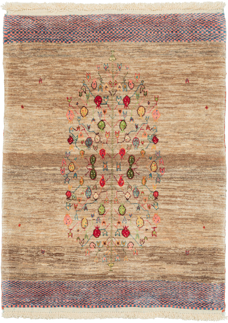 It features a a central tree of life motif with branches full of pomegranates in vibrant reds, blues, yellow and green. The tree sits atop a variegated field made up of various shades of undyed wool with a few small moments of color that add depth. Framed on top and bottom with a blue and pink checkered band. The pile is densely woven, making this a plush rug.