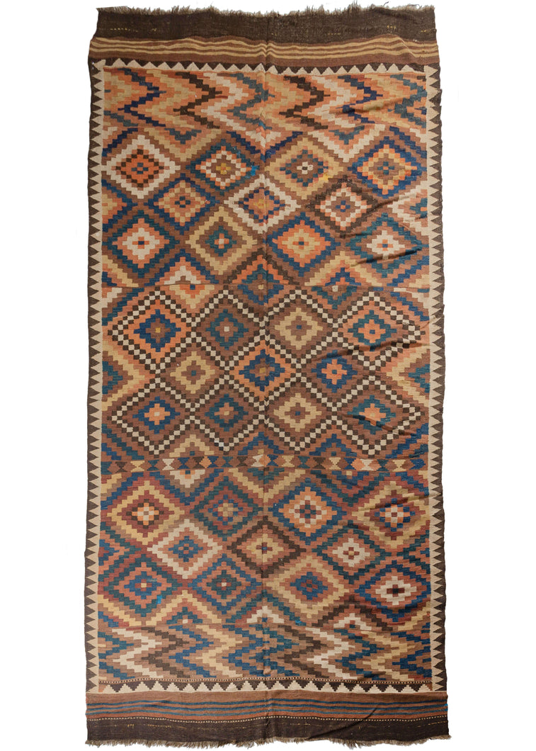 Antique Afghan Maimane kilim composed of a stepped-diamond pattern in light and dark blues, reds, browns, ivory, yellow, and orange. Multiple times the diamond pattern breaks and reforms into different variations of itself. Zig-zag sections reminiscent of lightning bolts add some electricity to each end and the whole is framed by a perimeter of reciprocating brown and white and brown triangles. The whole is finished with simple striped skirt kilim on either end.