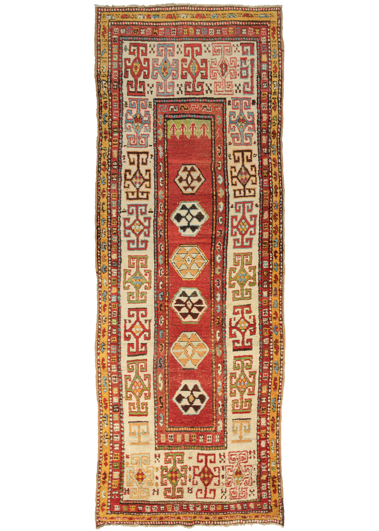 This Kazak Runner features a thin field of polygons on a red ground with arrows near a small green section at the top giving the pattern subtle directionality. The polygons are filled with protection symbols and slightly shift in scale unintentionally. It is framed by a very wide and relatively bright ivory main border with large-scale geometric devices in red, yellow, green, and icy blue.