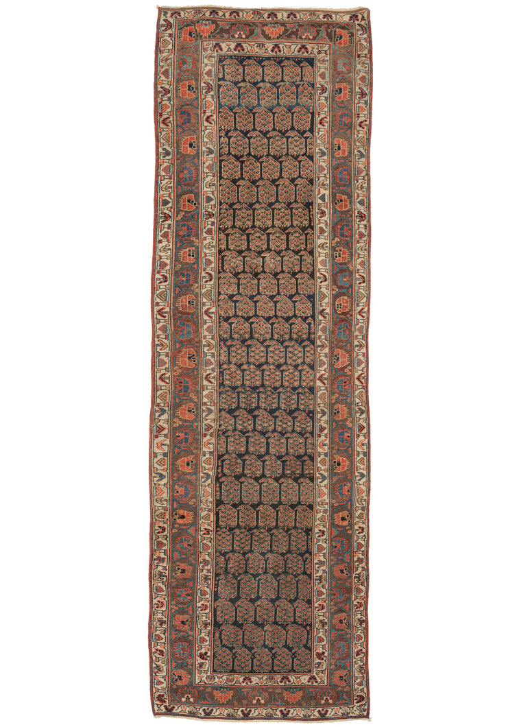 This Antique Kurdish Bidjar Runner features a field filled with offset rows of plump, polychrome botehs on a dark navy ground. The field is framed by a main border of scrolling vine work with alternating blossoms in attractive shades of red, blue, and yellow against a soft brown ground. It is flanked by two minor borders with a variation of the same pattern but less precise, on a smaller scale and in a perfectly contrasting ivory ground.