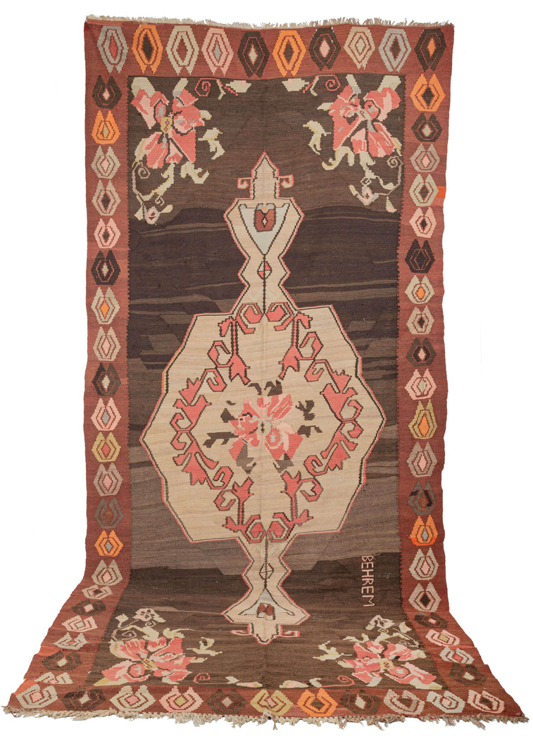 Turkish Kars Kilim featuring a scalloped central medallion with a dainty floral center surrounded by pink scrolls on camel ground. The same delicate flower featured in the medallion appears in each cornice. The field features a lovely brown which undulates wildly from light to dark brown which gives a subtle but pulsating energy. The word 