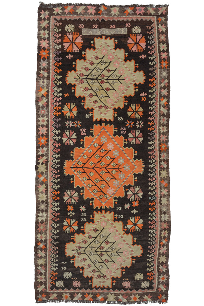 Mid century Caucasian kelleghi kilim featuring a triple medallion geometric design on a dark brown field. The medallions, alternating beige, and orange contain dainty flowers and leaves arranged in a diamond shape. The rest of the field is filled with various geometric shapes and symbols. At the top of the field are an inscription with numbers and letters reading: 